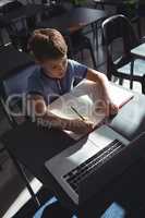 Boy writing in book by laptop at school