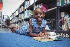 Smiling girl with book lying by shelf in library
