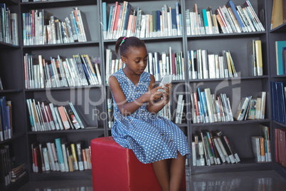 Girl using mobile phone while sitting against bookshelf in library