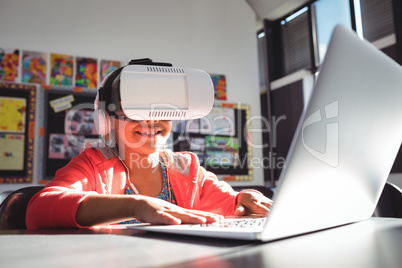 Smiling girl typing on laptop while using headphones and virtual reality glasses