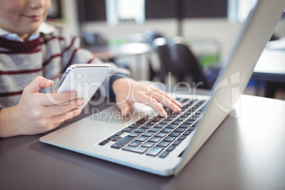 Mid section of schoolboy using laptop and mobile phone on desk