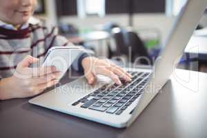 Mid section of schoolboy using laptop and mobile phone on desk