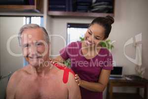 Smiling female therapist applying elastic therapeutic tape on shoulder of shirtless senior male pati
