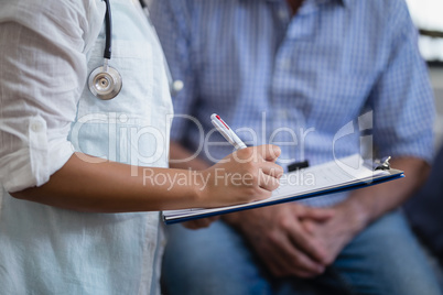 Midsection of female therapist writing on clipboard with male patient in background