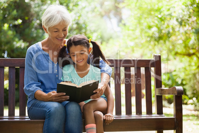 Smiling grandmother reading novel to granddaughter sitting on wooden bench