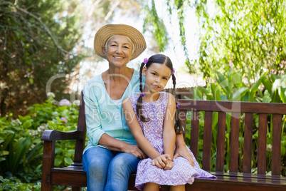 Portrait of senior woman wearing hat sitting with granddaughter on wooden bench