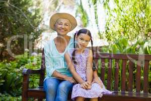 Portrait of senior woman wearing hat sitting with granddaughter on wooden bench