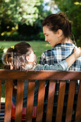 Smiling woman looking at daughter with arm around on wooden bench