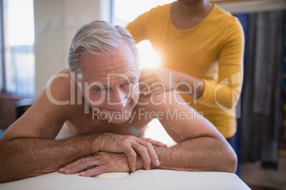 Smiling shirtless male patient lying on bed receiving neck massage from young female therapist
