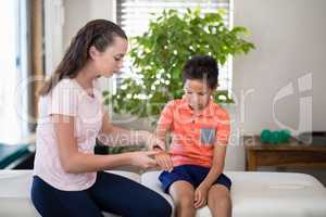 Young female therapist sitting on bed with boy examining hand