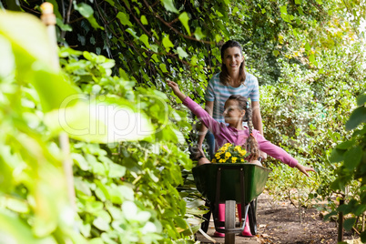 Smiling woman pushing girl sitting in wheelbarrow on footpath amidst plants