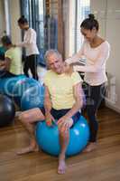 Smiling female therapist giving neck massage to senior patient sitting on exercise ball against mirr