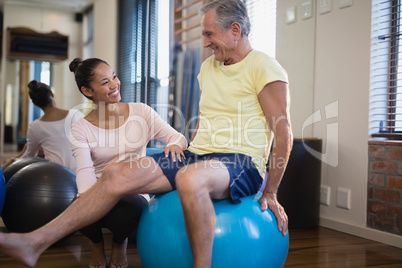 Smiling female therapist crouching by senior male patient sitting on exercise ball