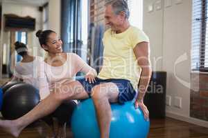 Smiling female therapist crouching by senior male patient sitting on exercise ball