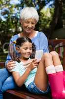 Smiling grandmother sitting with granddaughter using mobile phone on wooden bench
