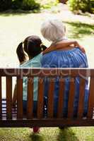 Rear view of girl with arm around grandmother sitting on wooden bench