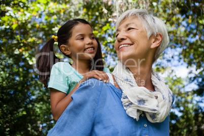 Low angle view of happy grandmother giving piggyback to granddaughter against trees