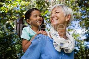 Low angle view of happy grandmother giving piggyback to granddaughter against trees