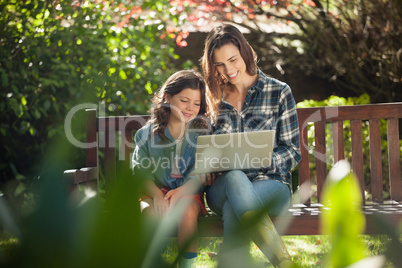 Smiling mother and daughter using laptop while sitting on wooden bench