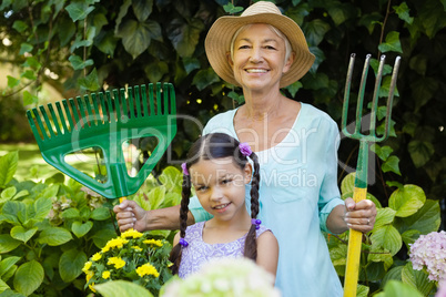 Portrait of smiling girl standing with grandmother holding gardening equipment