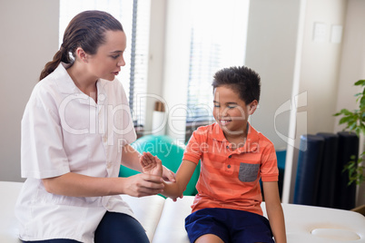 Young female therapist examining hand of smiling boy sitting on bed