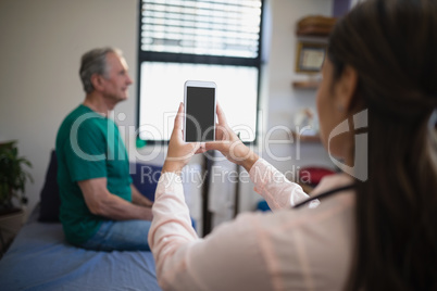Rear view of female therapist photographing senior male patient from mobile phone against window