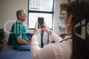 Rear view of female therapist photographing senior male patient from mobile phone against window