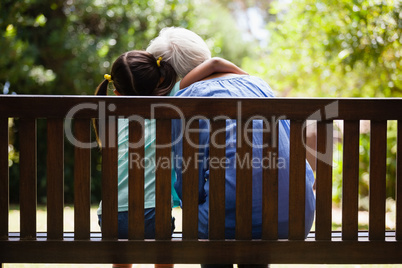 Rear view of granddaughter with arm around grandmother sitting on wooden bench