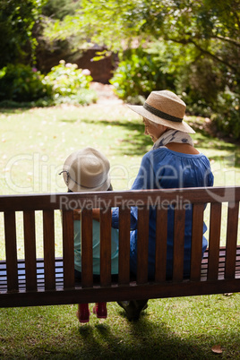 Rear view of granddaughter and grandmother wearing hats sitting on wooden bench