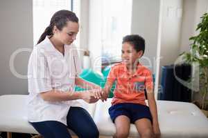 Female therapist examining hand of boy while sitting on bed