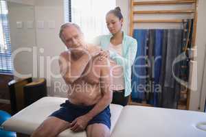 Shirtless senior male patient sitting on bed receiving neck massage from female therapist