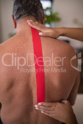 Hands of female therapist applying elastic therapeutic tape on shirtless senior male patient back