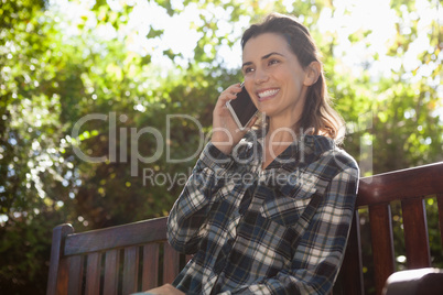 Low angle view of smiling beautiful woman talking on mobile phone while sitting on wooden bench