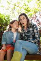 Girl whispering in ears of smiling surprised mother while sitting on bench