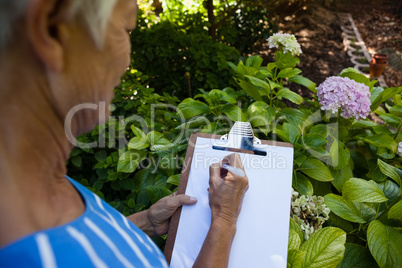 Midsection of senior woman writing on clipboard against plants