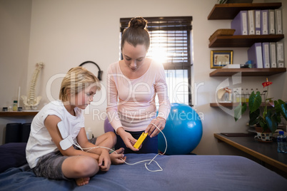 Female therapist showing electrical muscle stimulation machine to boy sitting on bed