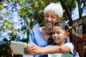 Smiling grandmother and granddaughter taking selfie while sitting on bench