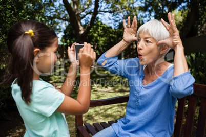Side view of girl taking photograph of grandmother making faces on bench