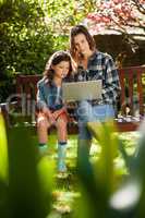 Girl sitting by mother using laptop on wooden bench