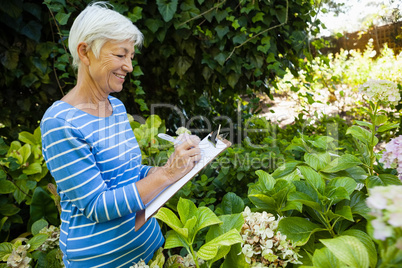 Smiling senior woman writing on clipboard while standing amidst plants