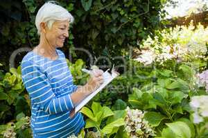 Smiling senior woman writing on clipboard while standing amidst plants
