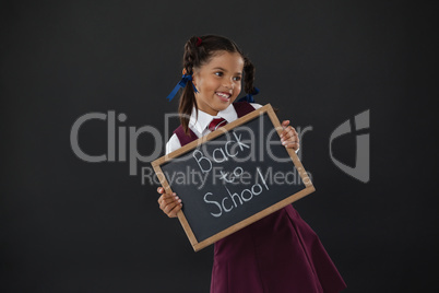 Smiling schoolgirl holding slate with text against blackboard