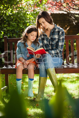 Mother reading book to daughter while sitting on wooden bench