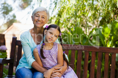 Portrait of smiling senior woman sitting with arm around girl on wooden bench
