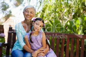 Portrait of smiling senior woman sitting with arm around girl on wooden bench