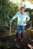Smiling senior woman wearing hat standing with garden fork on dirt