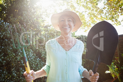 Back lit portrait of smiling senior woman holding garden fork and shovel