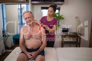 Female therapist applying elastic therapeutic tape on shoulder of frowning shirtless senior male pat