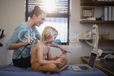 Smiling female therapist pointing at laptop while scanning shoulder of shirtless boy