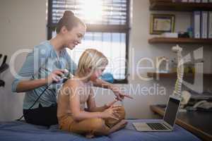 Smiling female therapist pointing at laptop while scanning shoulder of shirtless boy